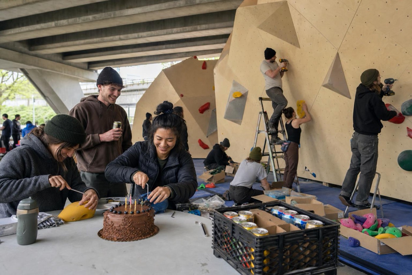 Freiwillige bauen eine Boulderwand, vorne wird Kuchen geschnitten und Griffe verschraubt